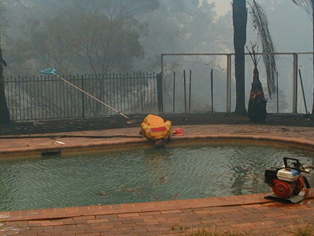 A firefighter drafting water from a backyard swimming pool with a portable pump, smoke-filled bush behind the fence