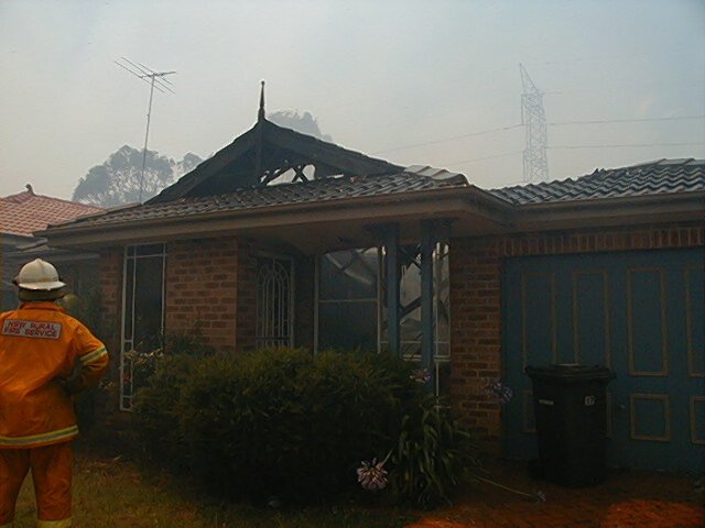 A suburban brick house with its roof burnt through, a rural fire service firefighter standing in front