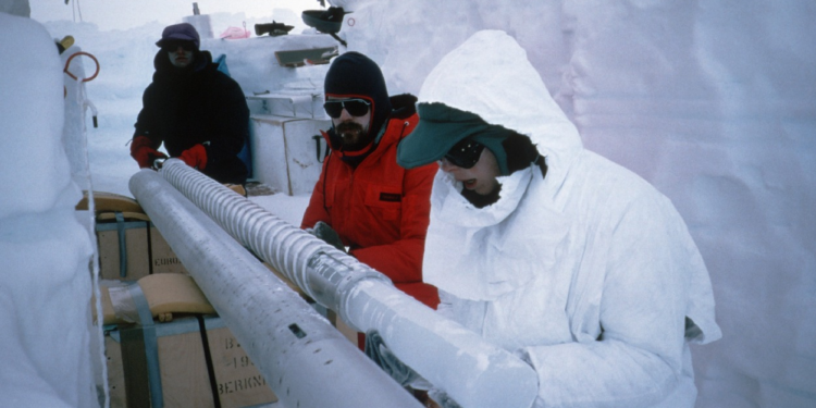 Researchers in protective cold-weather clothing handling a long cylindrical ice core sample at a polar drilling site